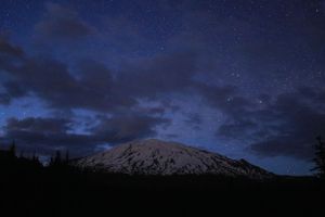 Snow-capped mountain silhouetted against starry night sky with dark forest in foreground.