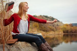Person in red cardigan sitting by lake during autumn with arms outstretched.