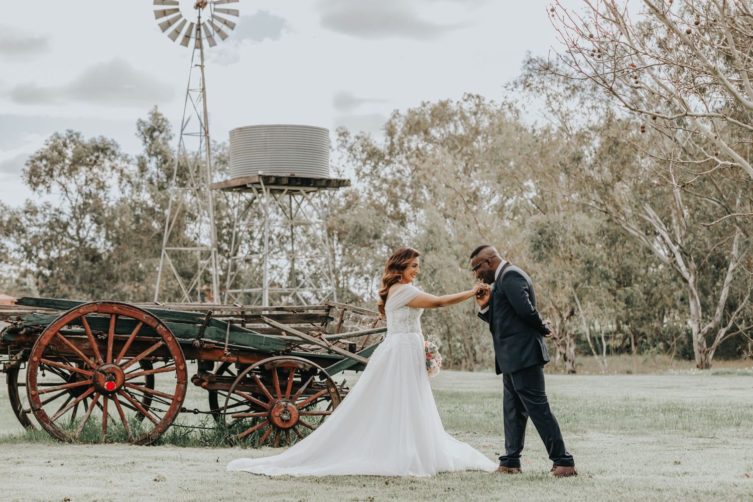 Wedding couple dances near rustic wagon and windmill in ethereal countryside setting.