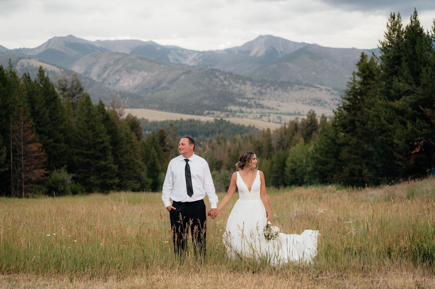 Wedding couple holding hands in a golden meadow with mountain and pine forest backdrop.