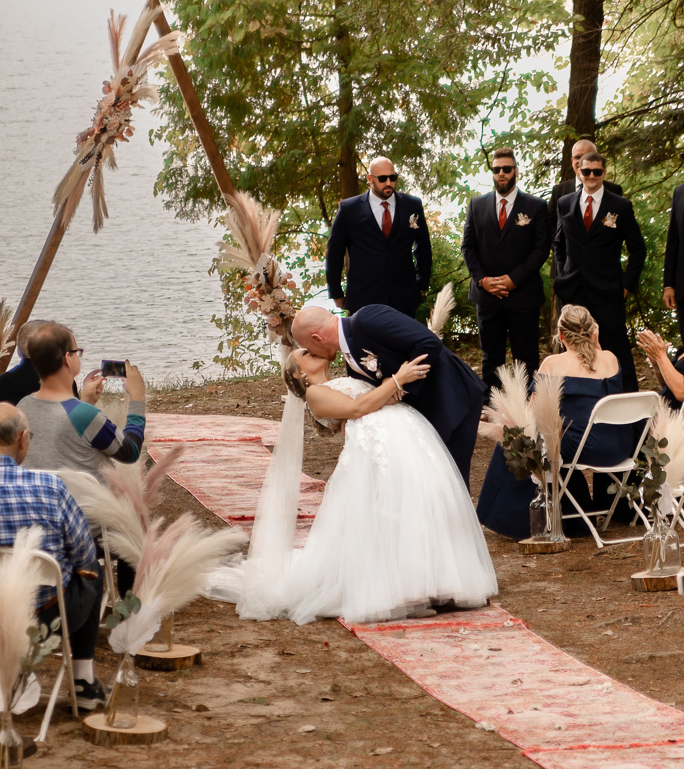 Wedding ceremony setup on a lakeside beach with folding chairs and decorative rugs.