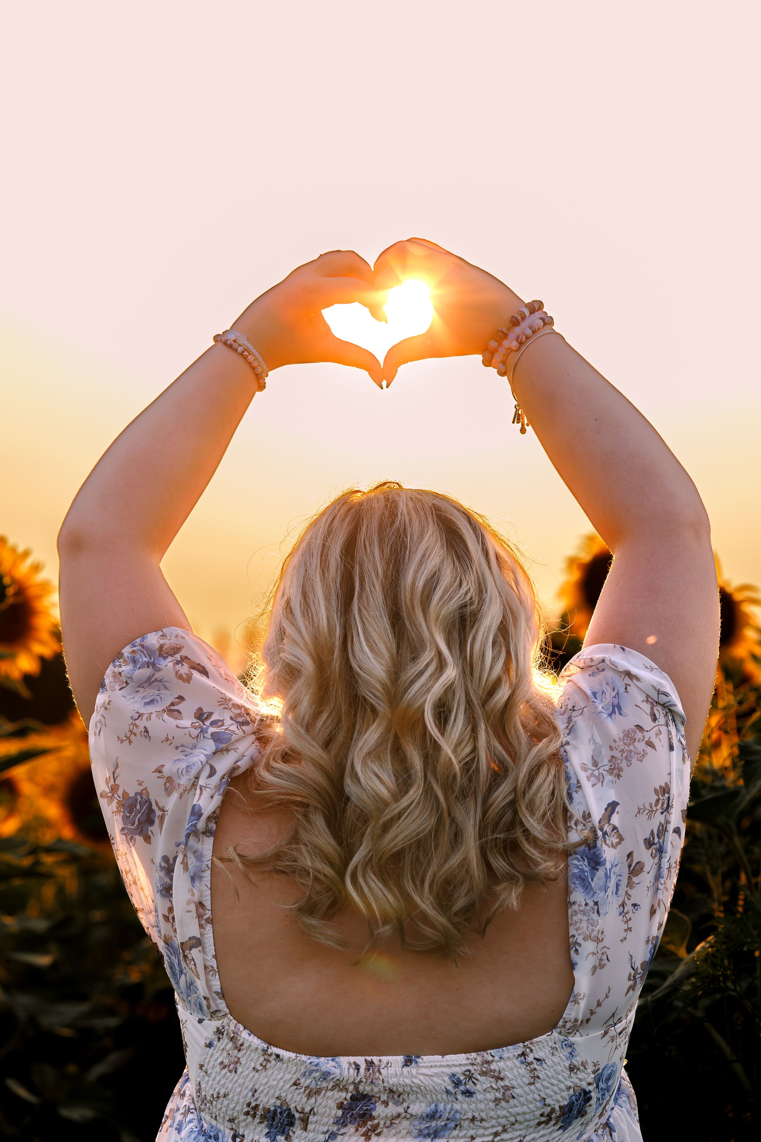 Woman in floral dress makes heart shape with hands against sunset sky in romantic silhouette.