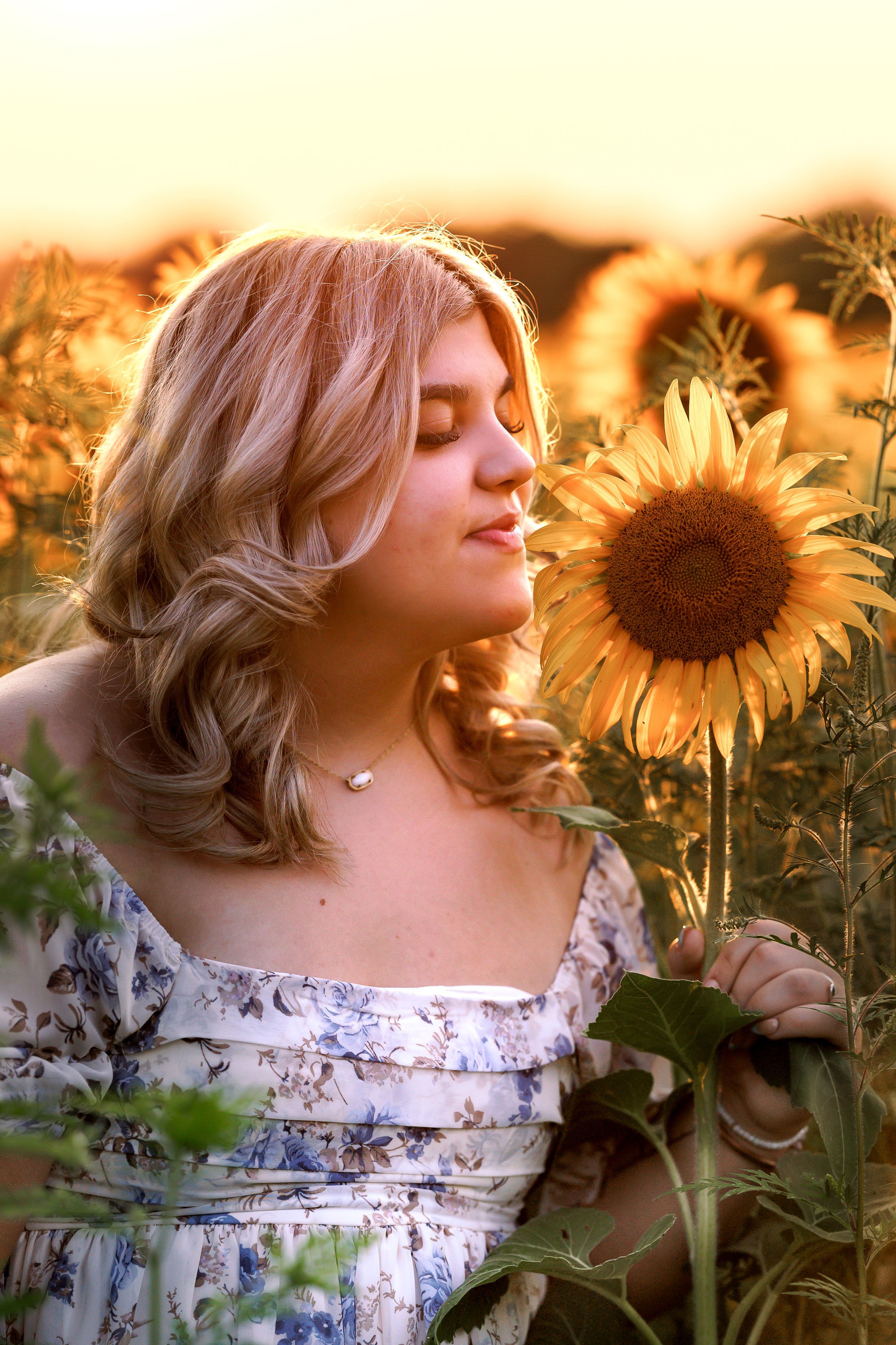 Woman in floral dress poses dreamily among blooming sunflowers at golden hour.