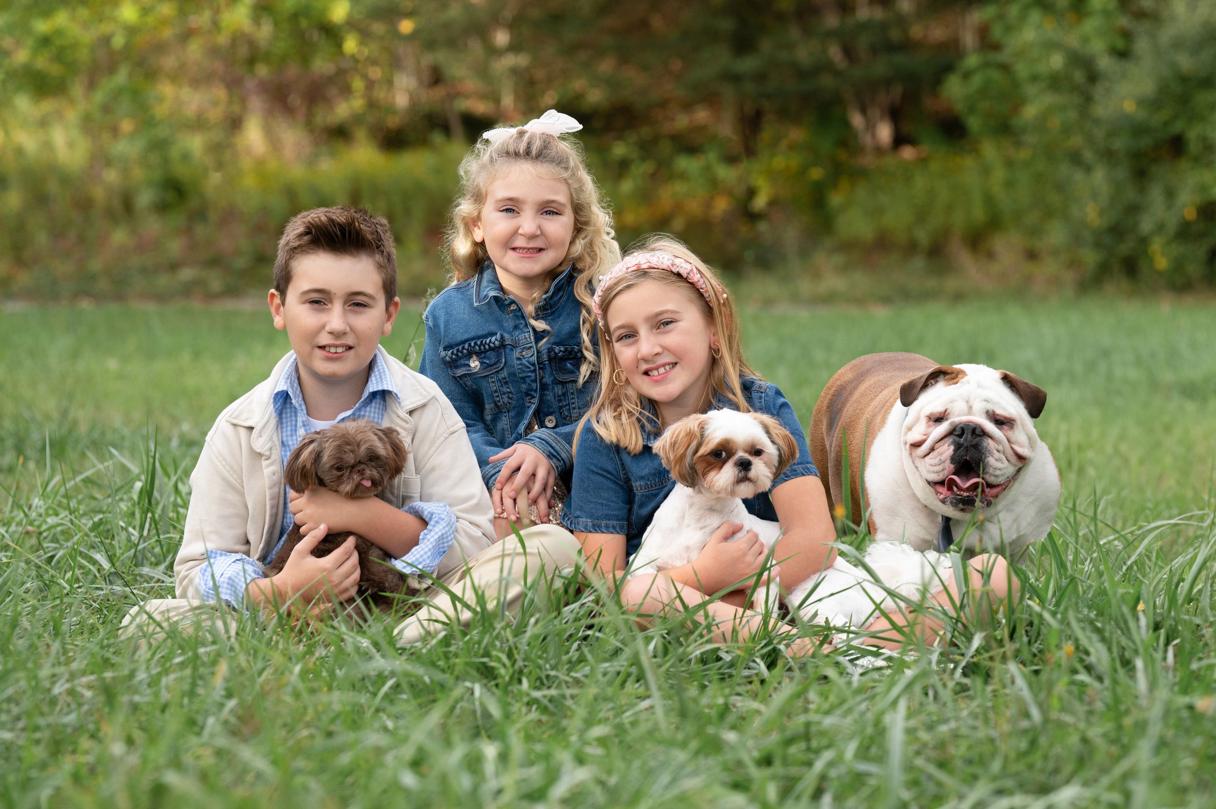 Three friends sit in tall grass with their pets including puppies and a pug dog on a sunny day outdoors.