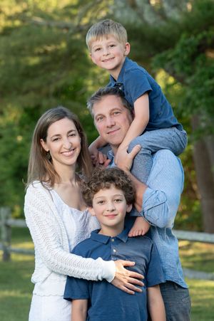 A family of four poses together outdoors on a sunny day with greenery in the background.