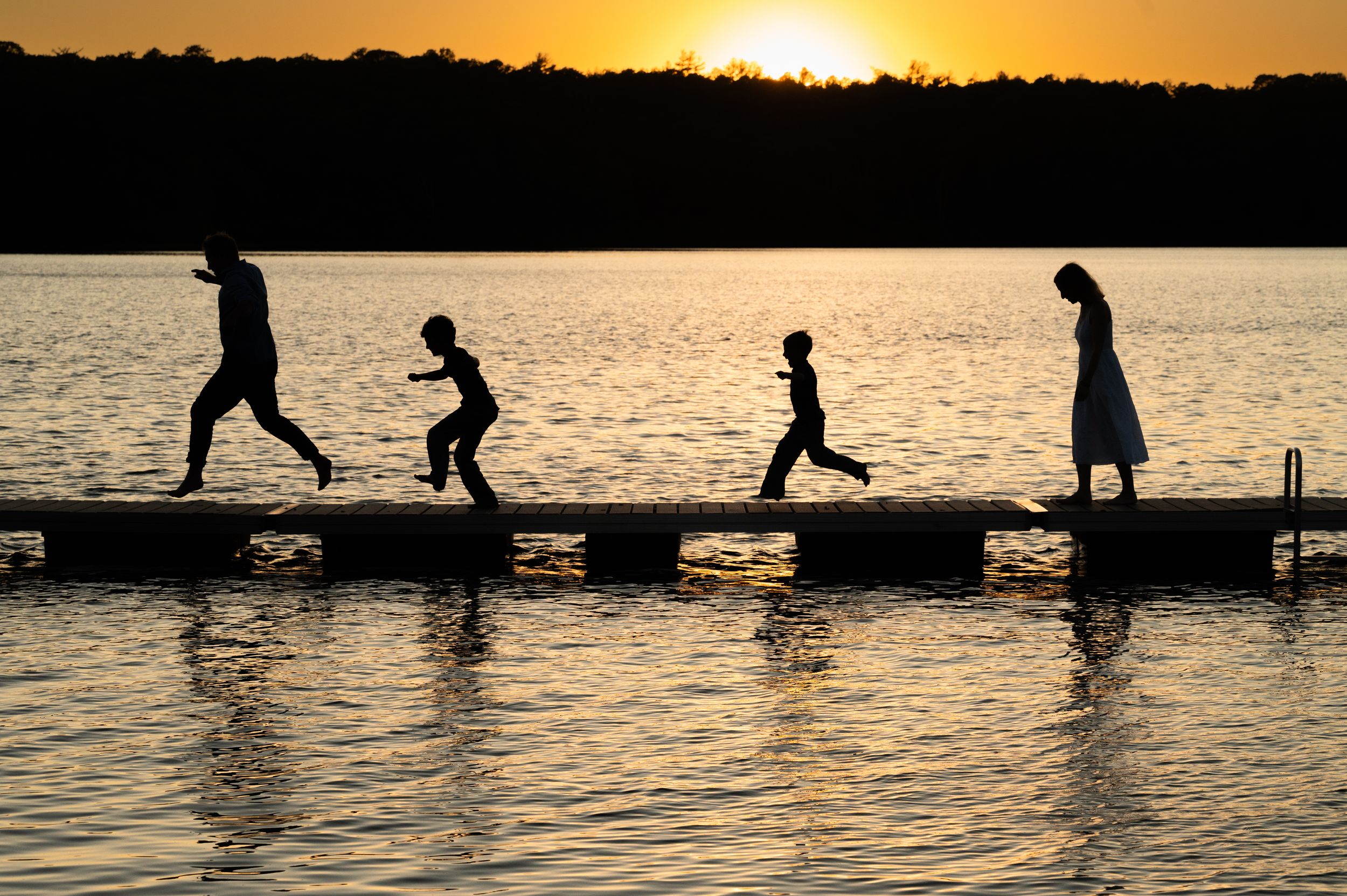 Silhouettes play and leap across a wooden dock at sunset over a serene lake.