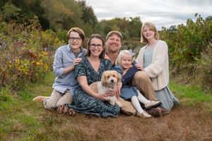 A family sits together outdoors in a natural setting with wildflowers, accompanied by their golden retriever dog.