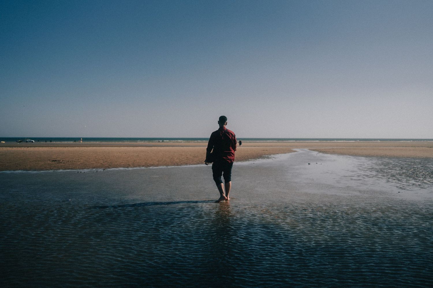 Silhouette walking alone on beach during low tide against clear blue sky.