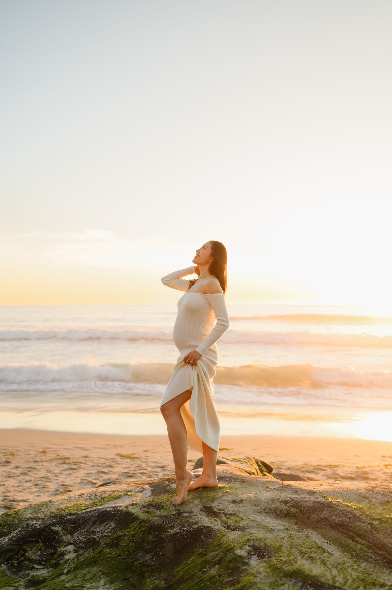 woman in knit dress stands on beach rocks in front of waves during her sunset beach photoshoot by Ventura and Santa Barbara Photographer
