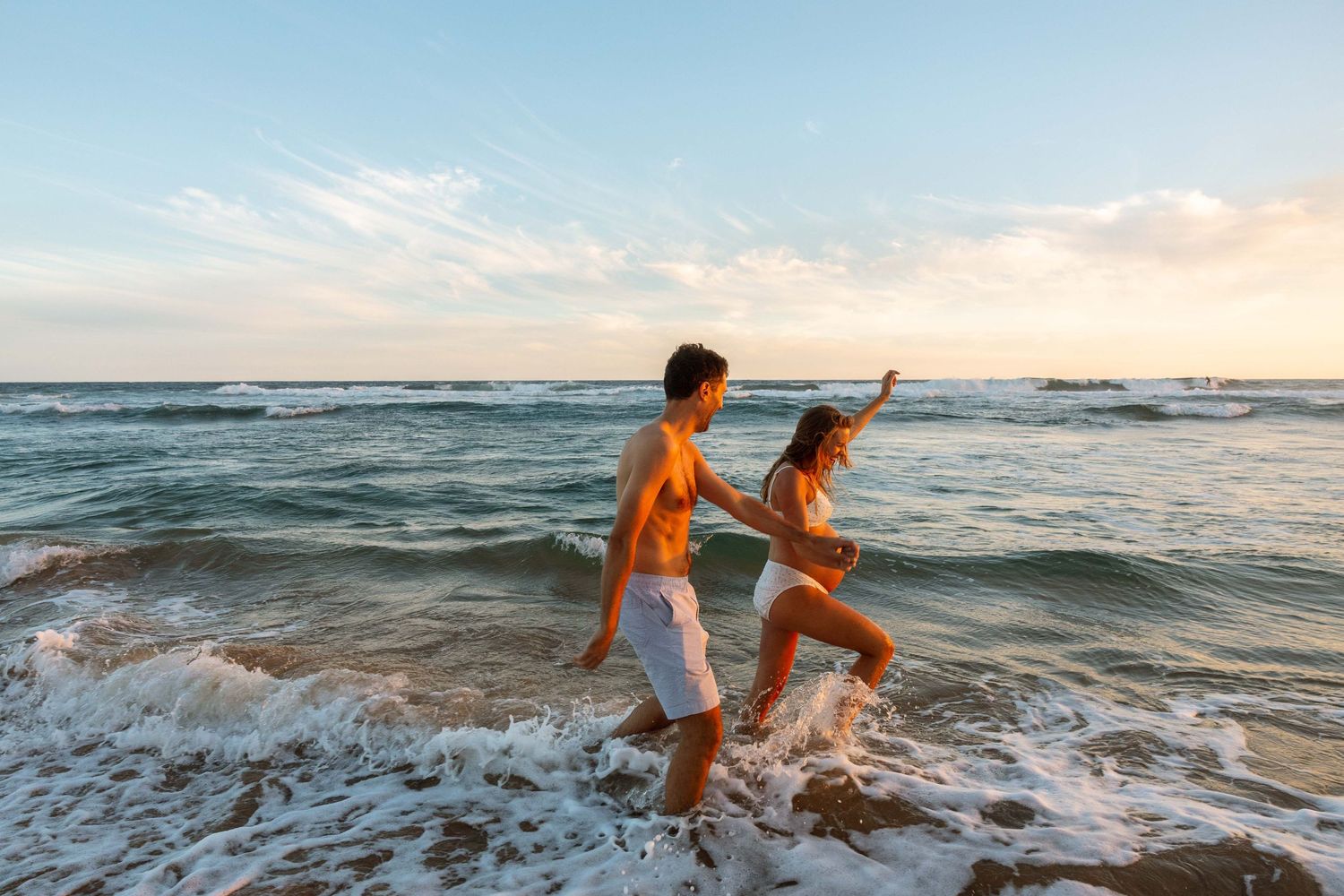 A beautiful young couple playing at the beach in the waves, while expecting their first child.