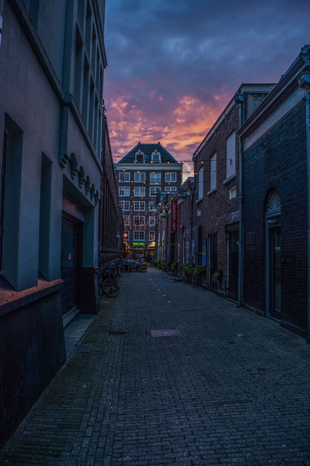Moody sunset over narrow Amsterdam alley with historic buildings and cobblestone street.