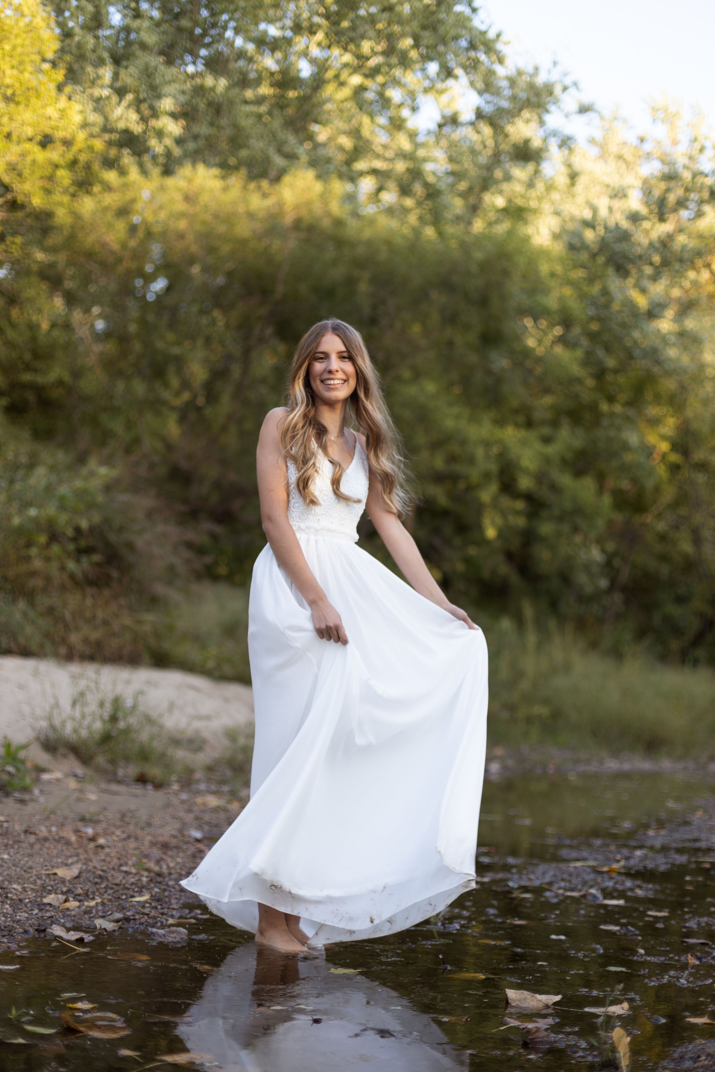 High school girl kicking up creek water while wearing an iconic floor length white dress.
