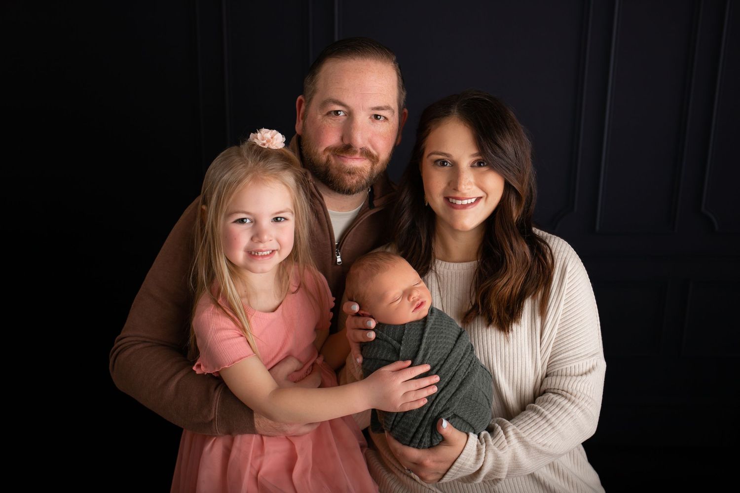 Family portrait with parents, young child and newborn baby against dark background.