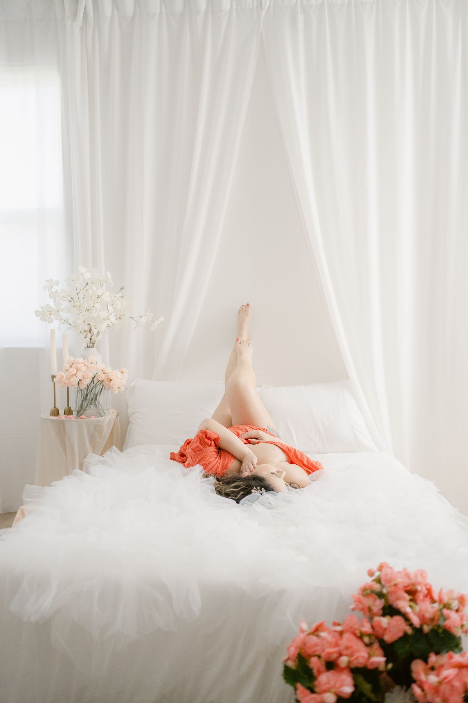 A dreamy boudoir photo with soft white curtains and coral flowers featuring draped fabrics on white bedding.