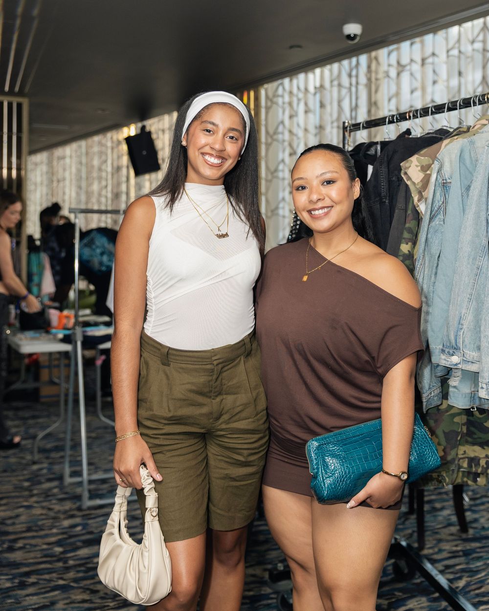Two smiling women pose together at a fashion pop-up event, one holding a white bag, the other a teal clutch.