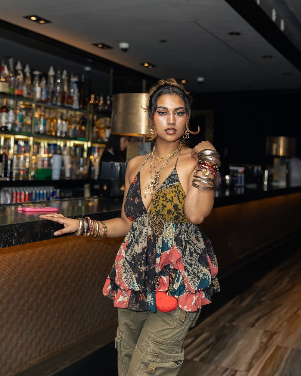 A stylish woman in a floral top and cargo pants poses at a bar holding a drink, adorned with gold jewelry.