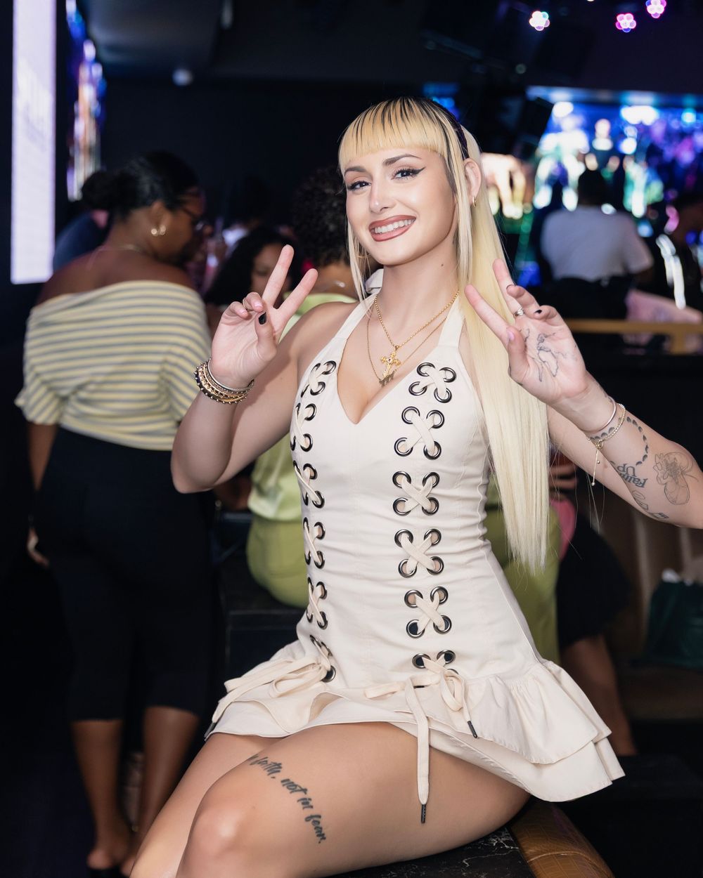 Blonde woman in white lace-up mini dress posing with peace signs at a nightclub party.