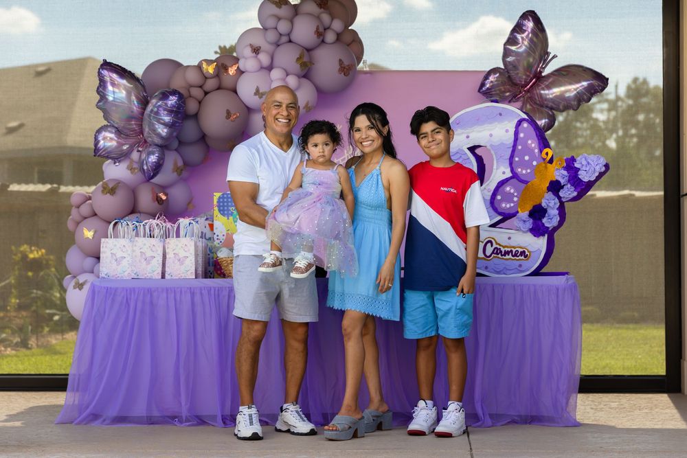 Smiling family of four poses beside a purple butterfly-themed birthday party table with balloons, a toddler in a tutu, and gift bags.