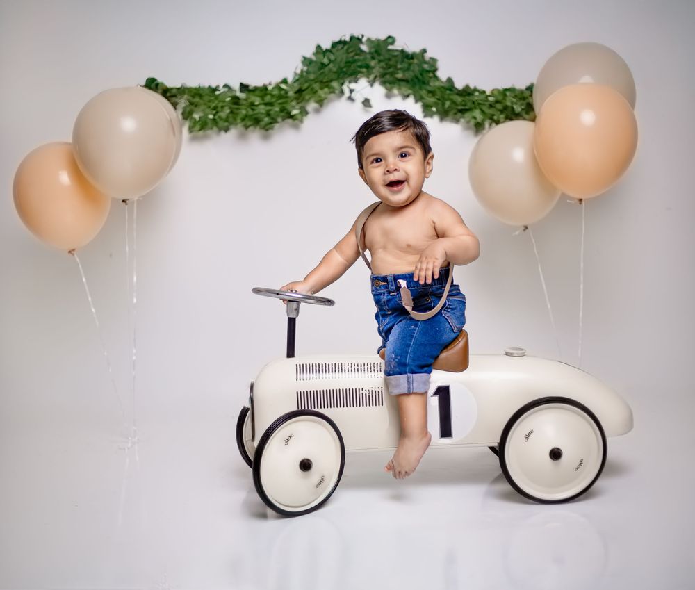 A joyful baby in blue overalls stands on a vintage white toy car with peach balloons and greenery decorations in the background.