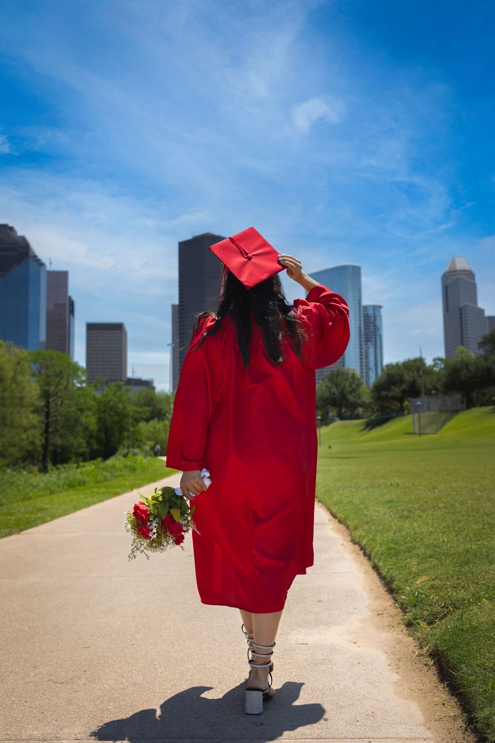 Graduate in red cap and gown holding flowers stands on path with city skyline and green park in background.