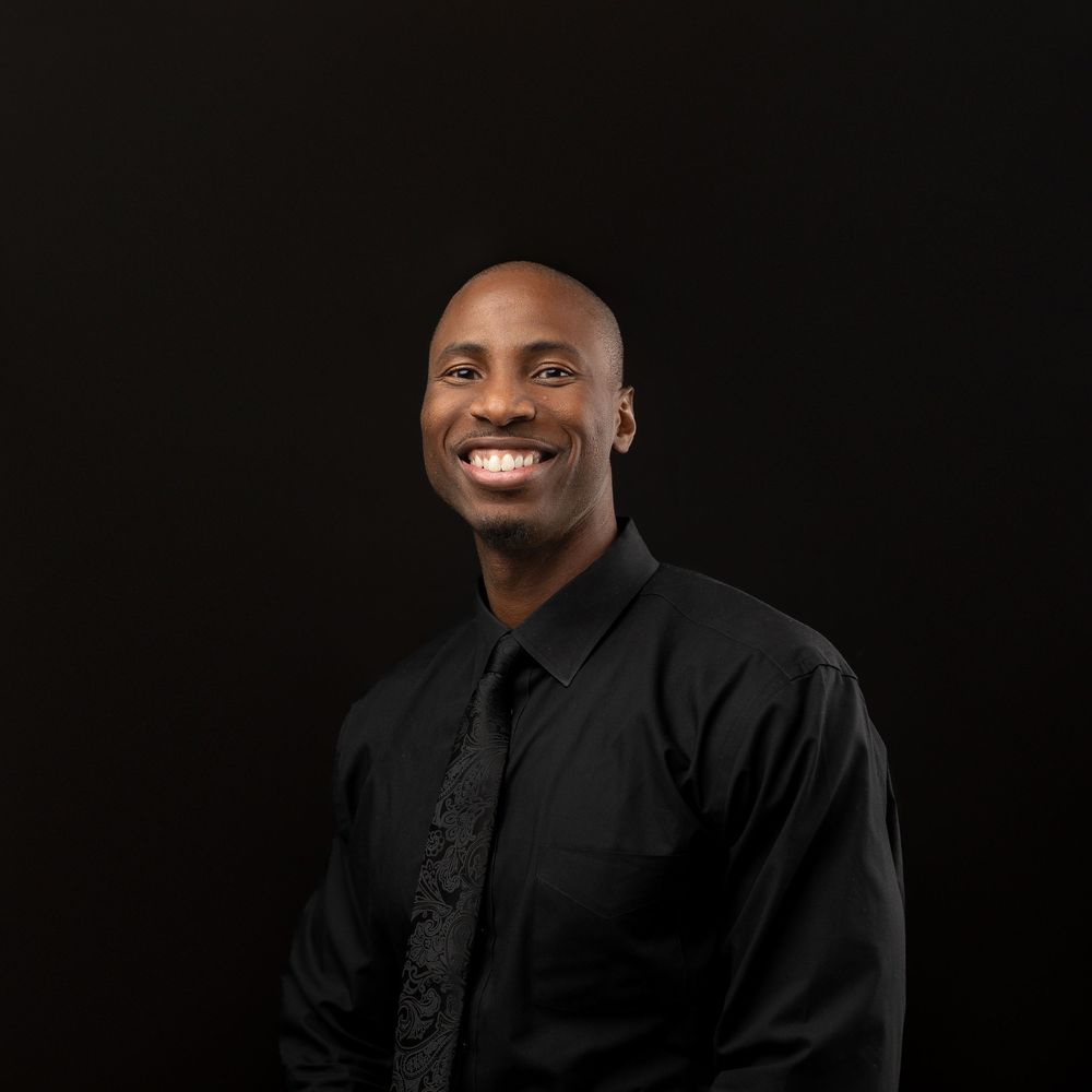 Professional studio portrait of a person wearing a black dress shirt and tie smiling against a dark background.