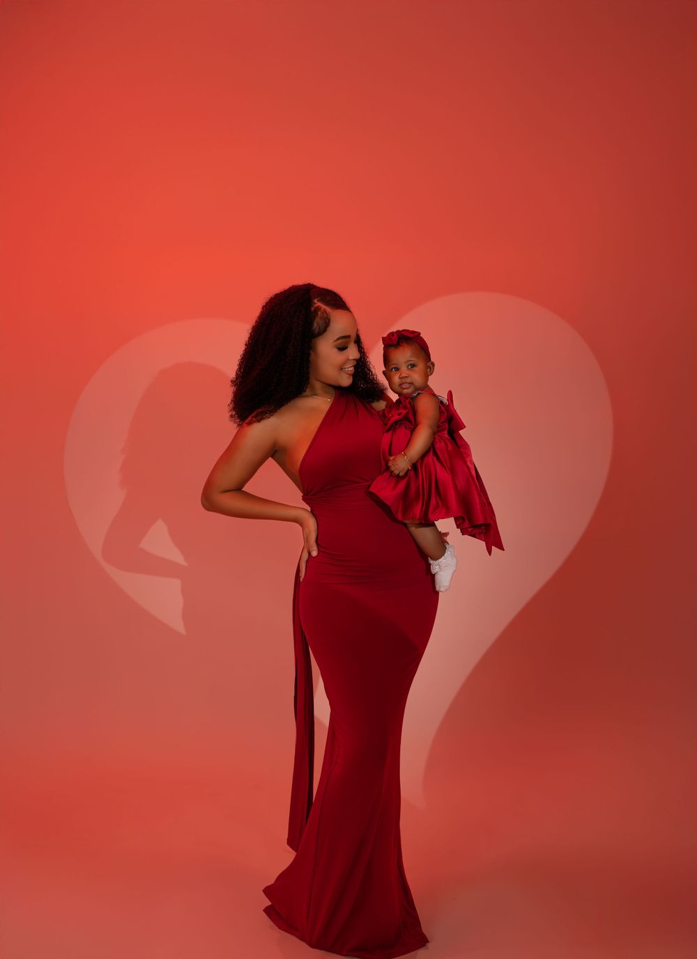 A figure in a red floor-length dress poses with a small child wearing a matching red outfit against a red background.