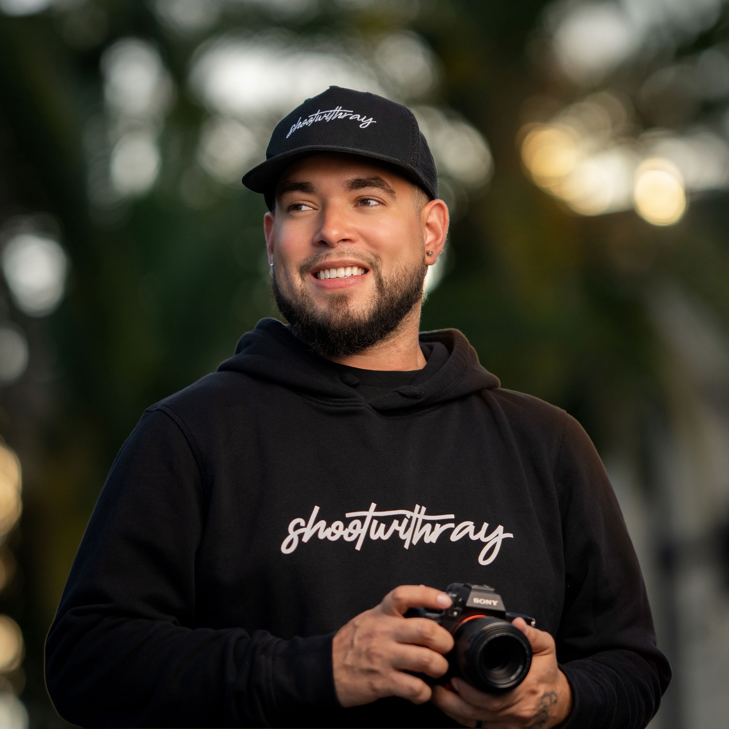 A photographer in a brown hoodie holds a DSLR camera against an orange-lit background, smiling warmly.