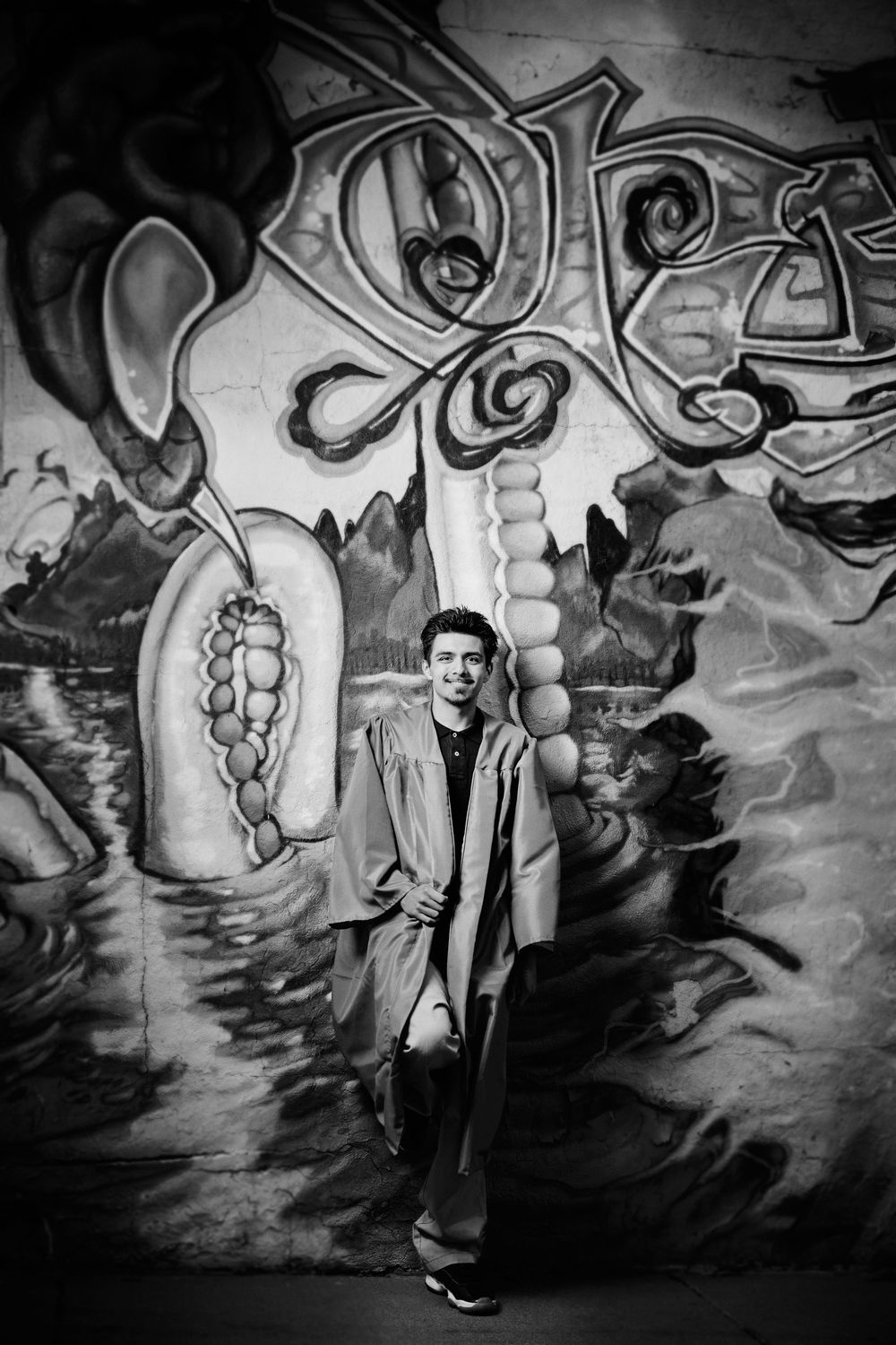 Black and white photograph of someone posing in front of artistic graffiti mural with abstract swirling designs.