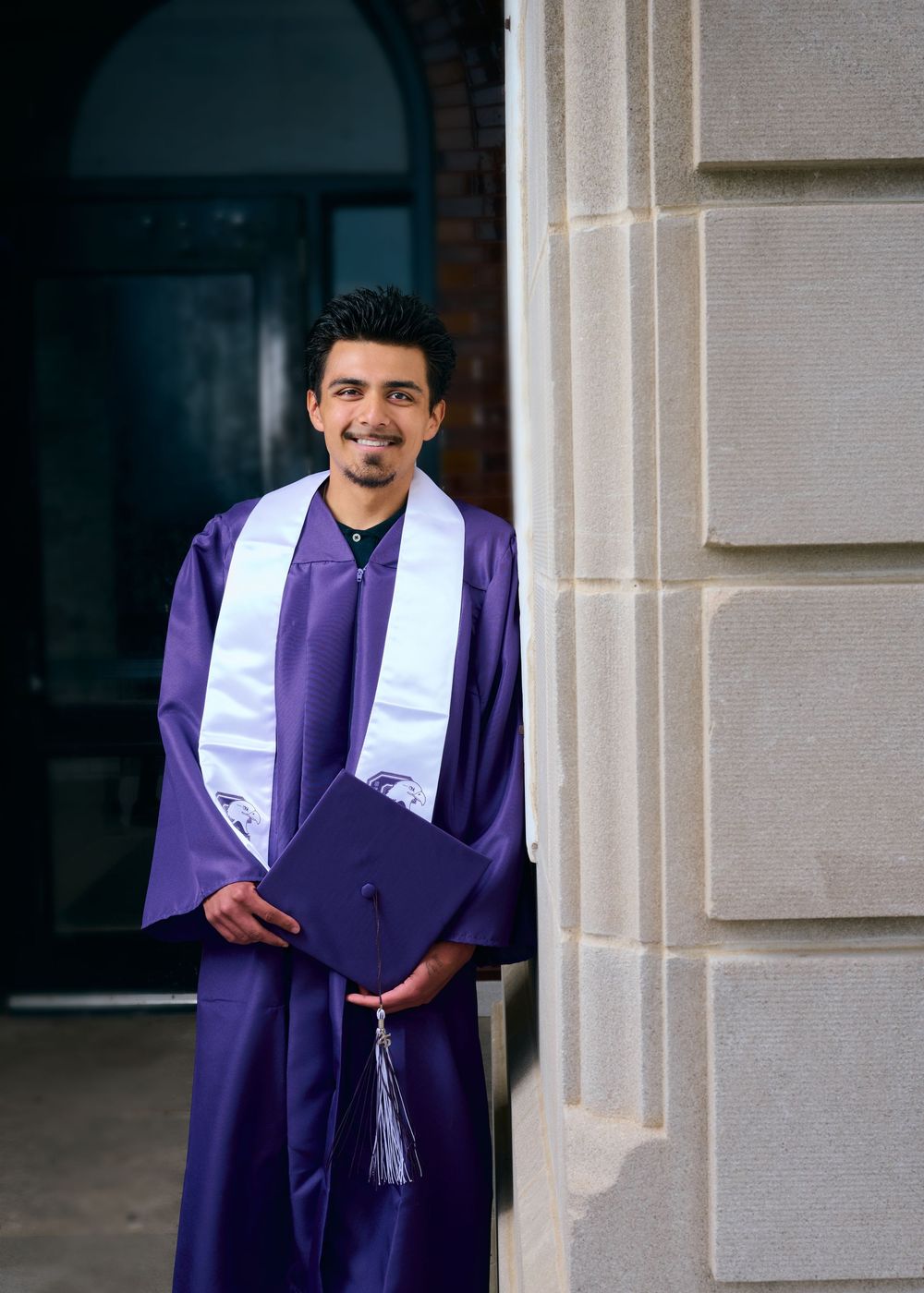 Graduate in purple cap and gown smiling while standing next to stone column.
