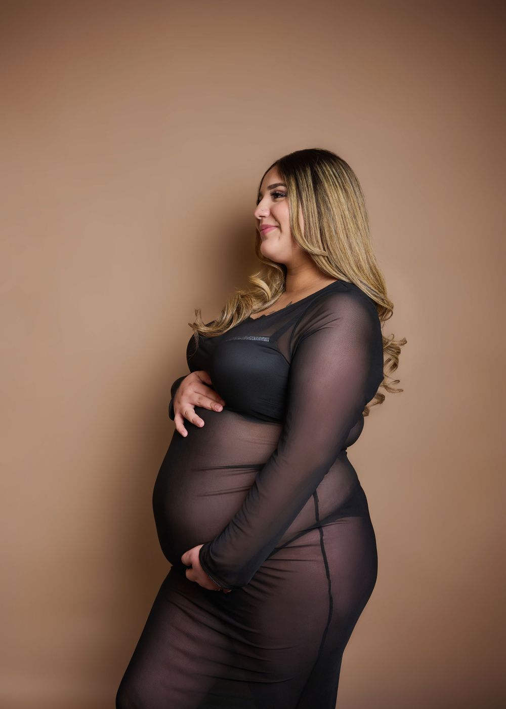 Side view maternity portrait in sheer black dress against neutral backdrop.