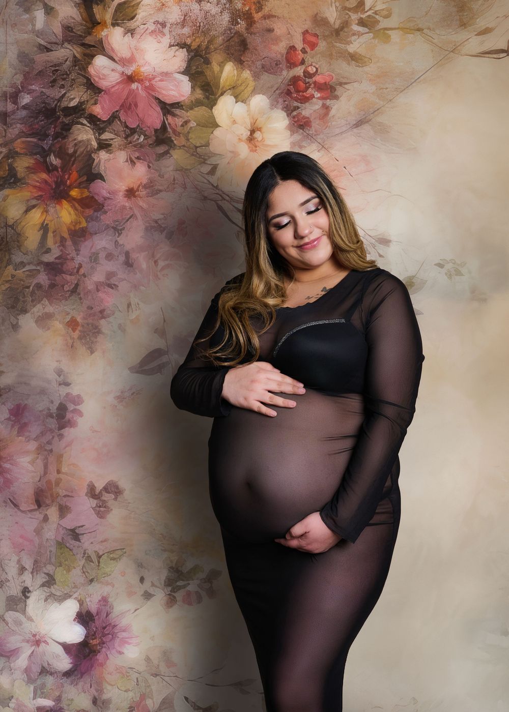 A pregnant person in a sheer black dress poses against a floral backdrop during a maternity photoshoot.