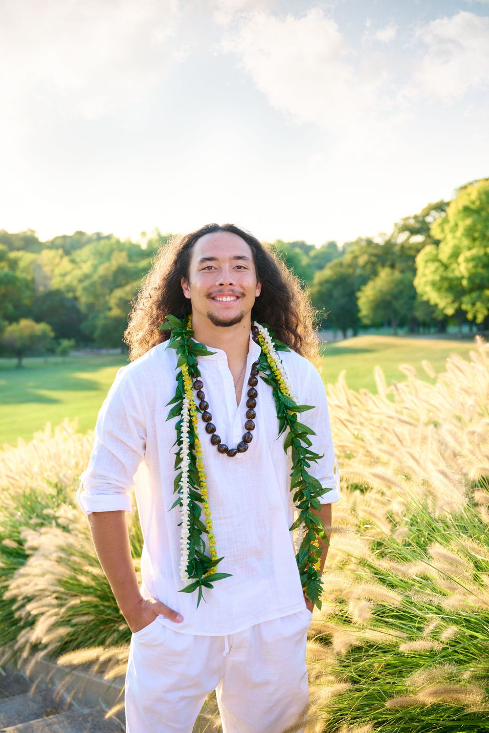 Person in white attire wearing green lei poses in sunny outdoor setting.