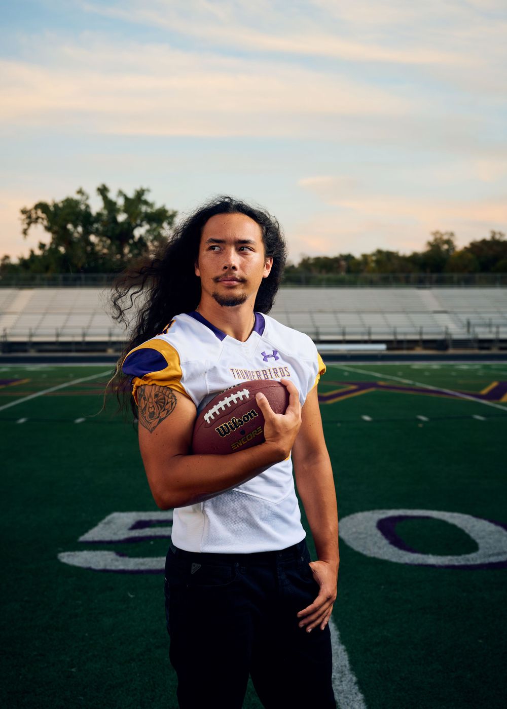Football player poses with ball on field wearing white jersey at sunset.