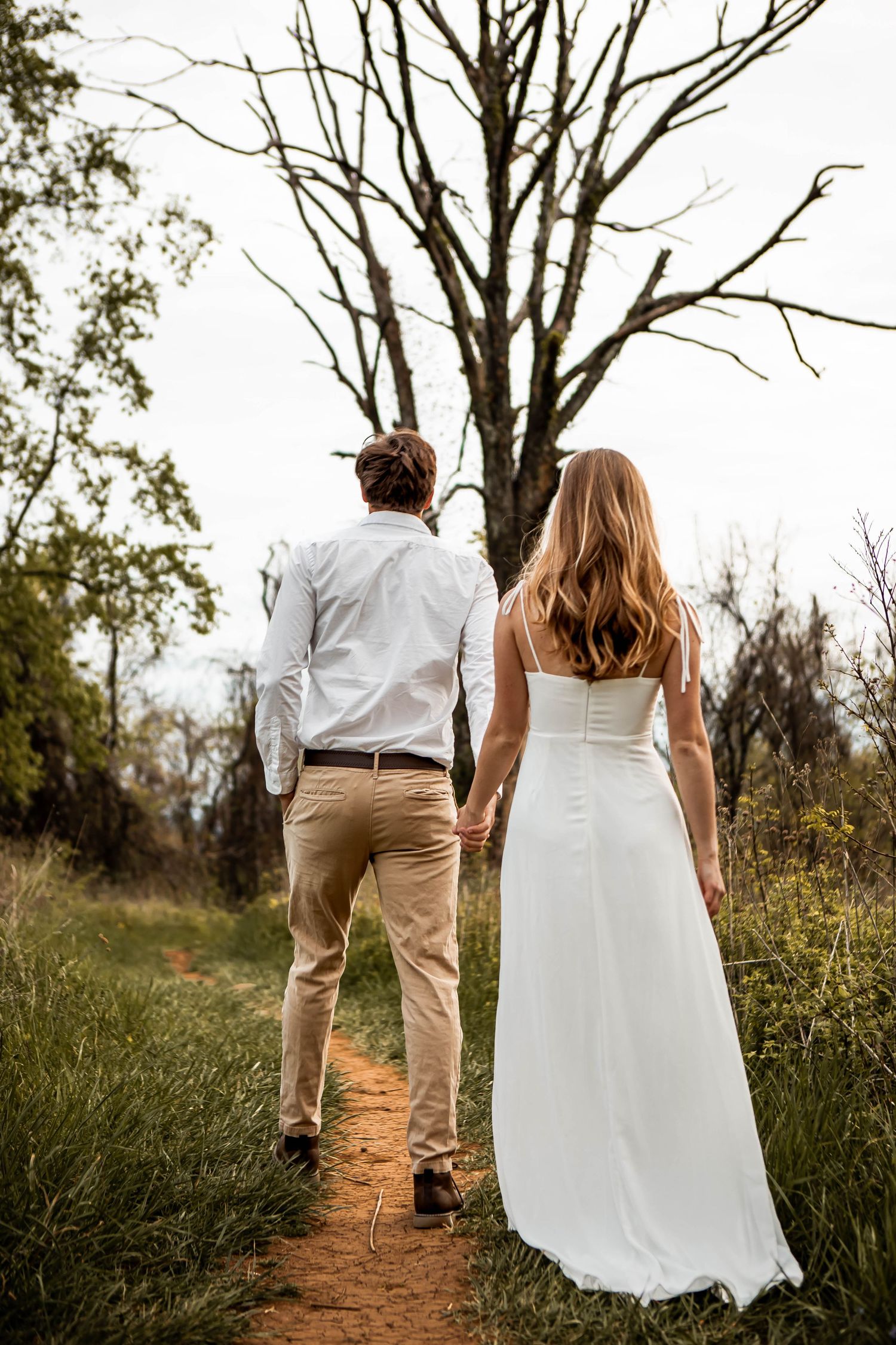 Couple walking down dirt path together surrounded by tall trees and natural scenery.