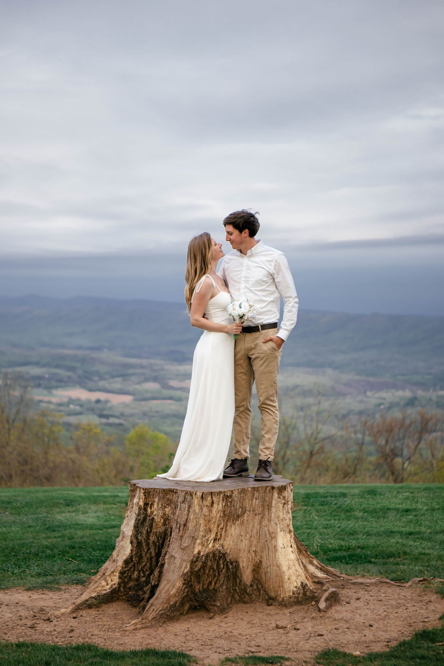 A couple in wedding attire stands intimately on a tree stump overlooking a mountain landscape.