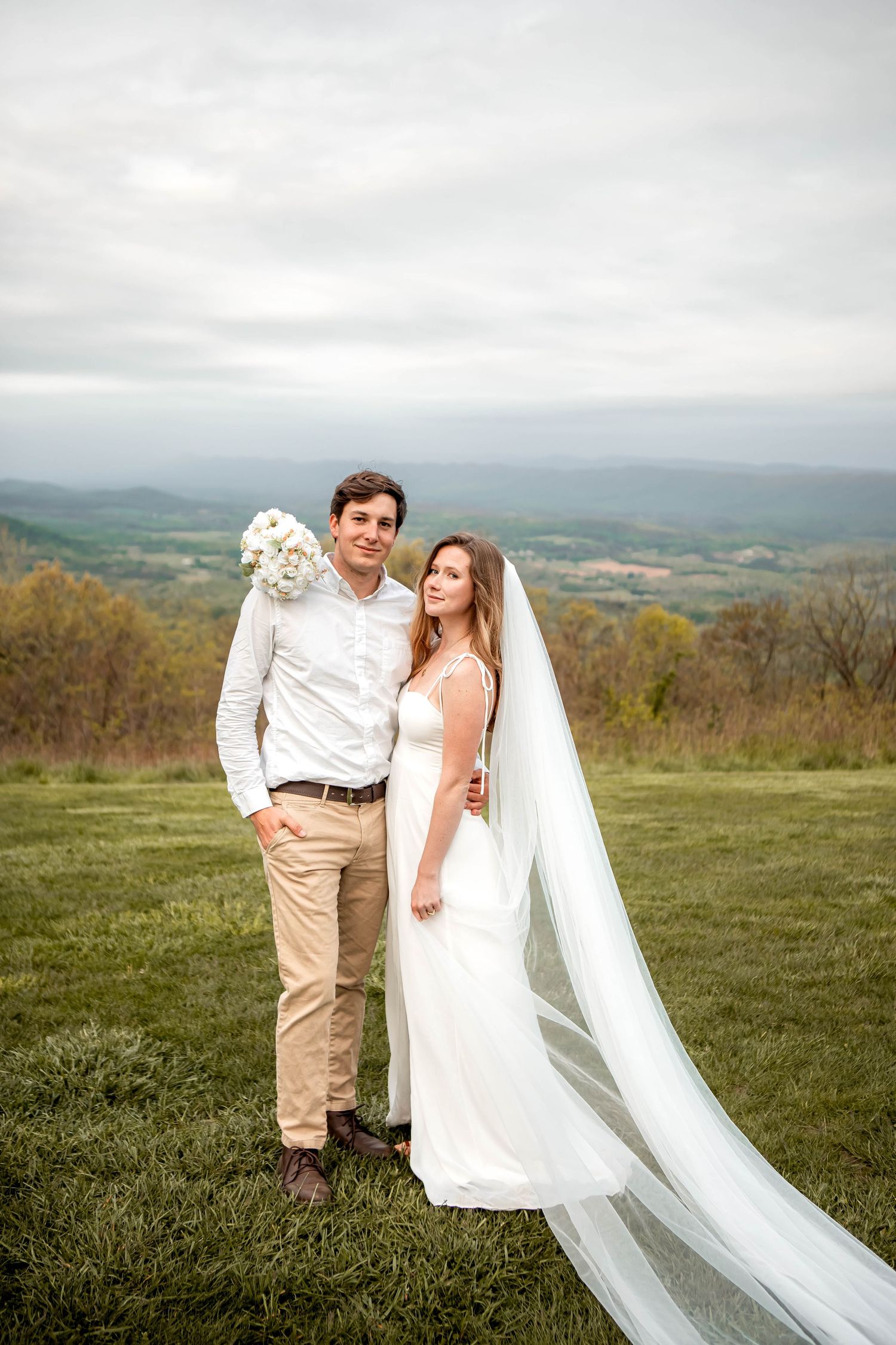 A couple in wedding attire poses together on a hilltop meadow with a scenic mountain landscape in the background.