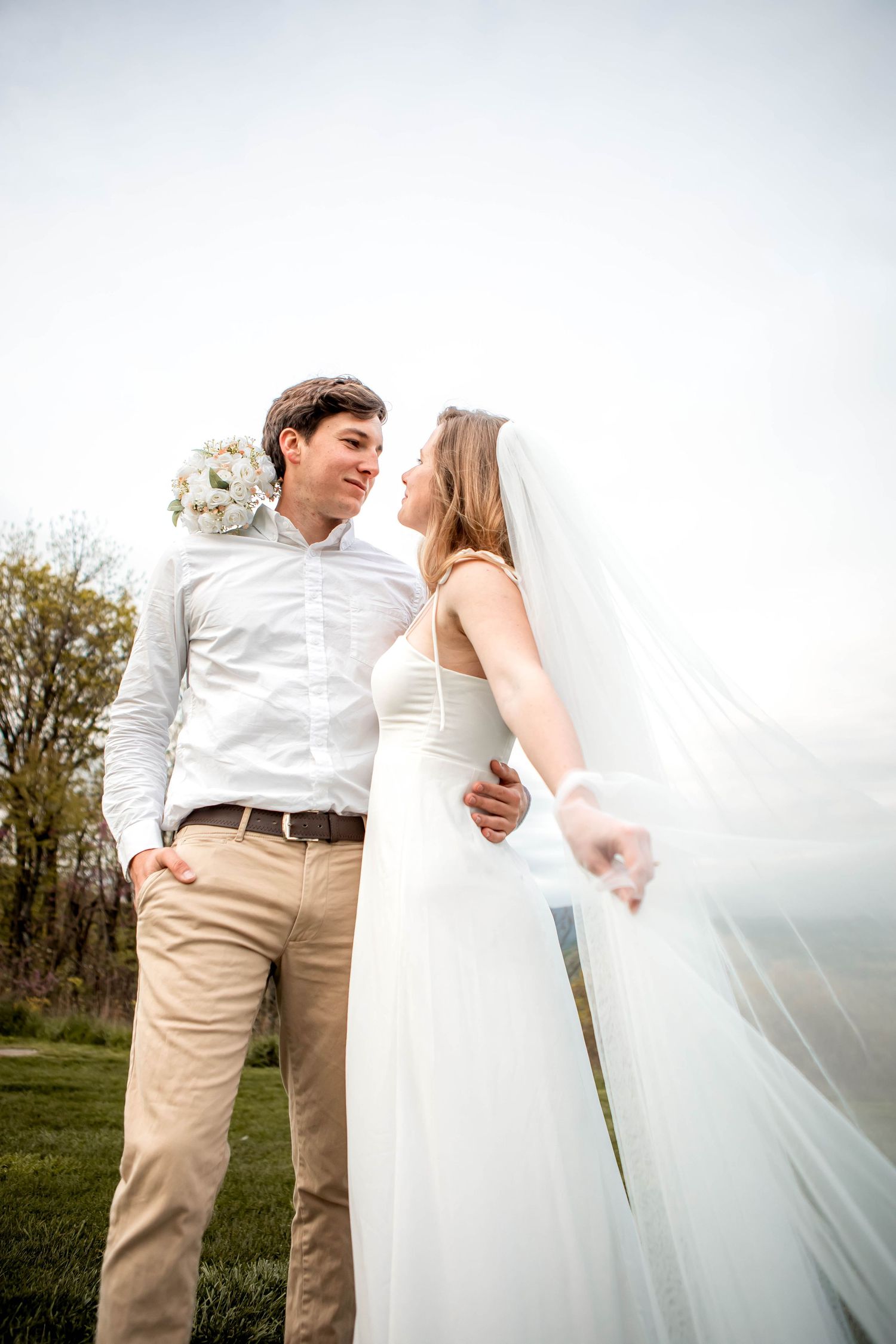 A couple shares an intimate moment in wedding attire against a bright white background with flowing veil.