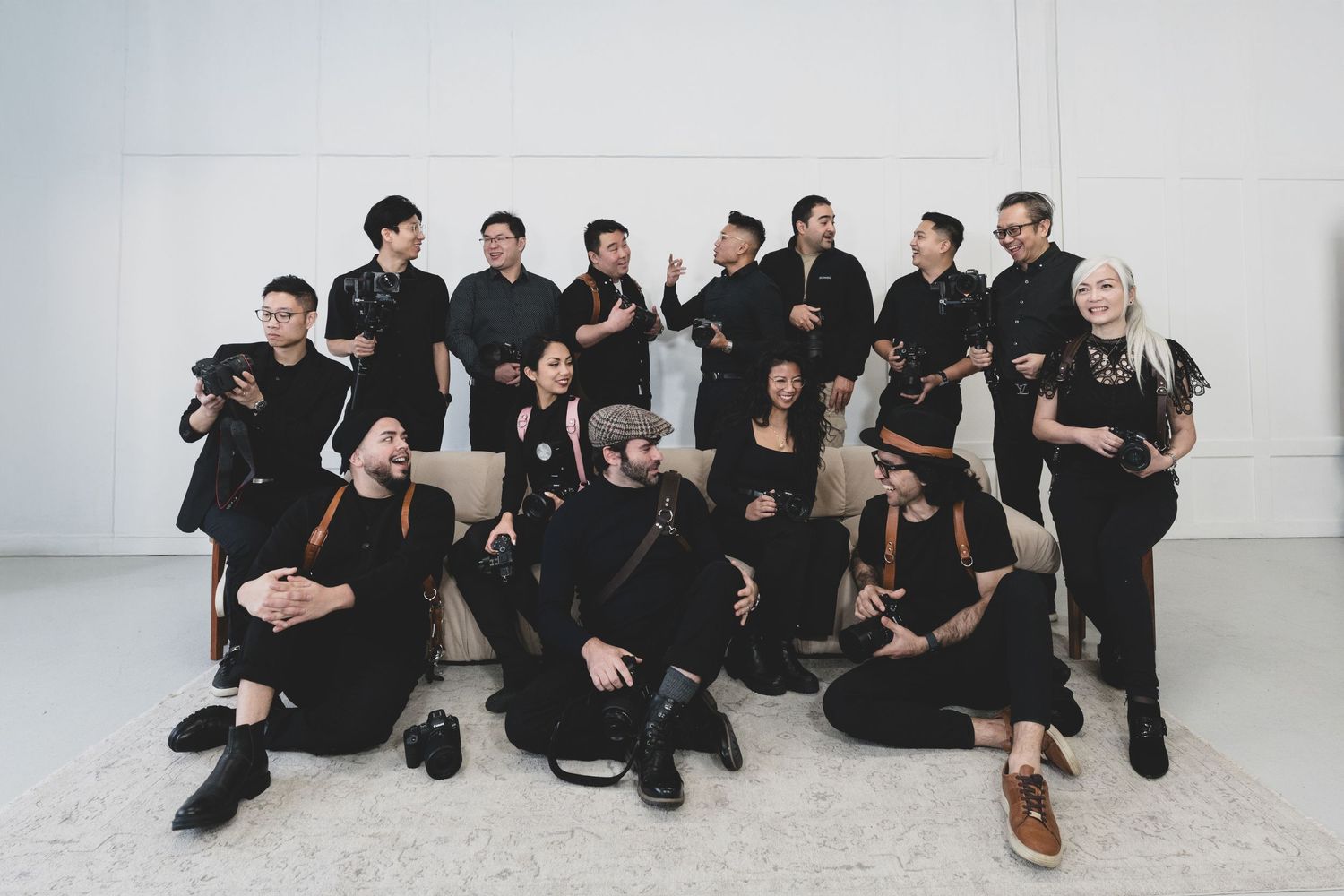 A group of individuals wearing all black outfits pose together against a white wall in a minimalist setting.