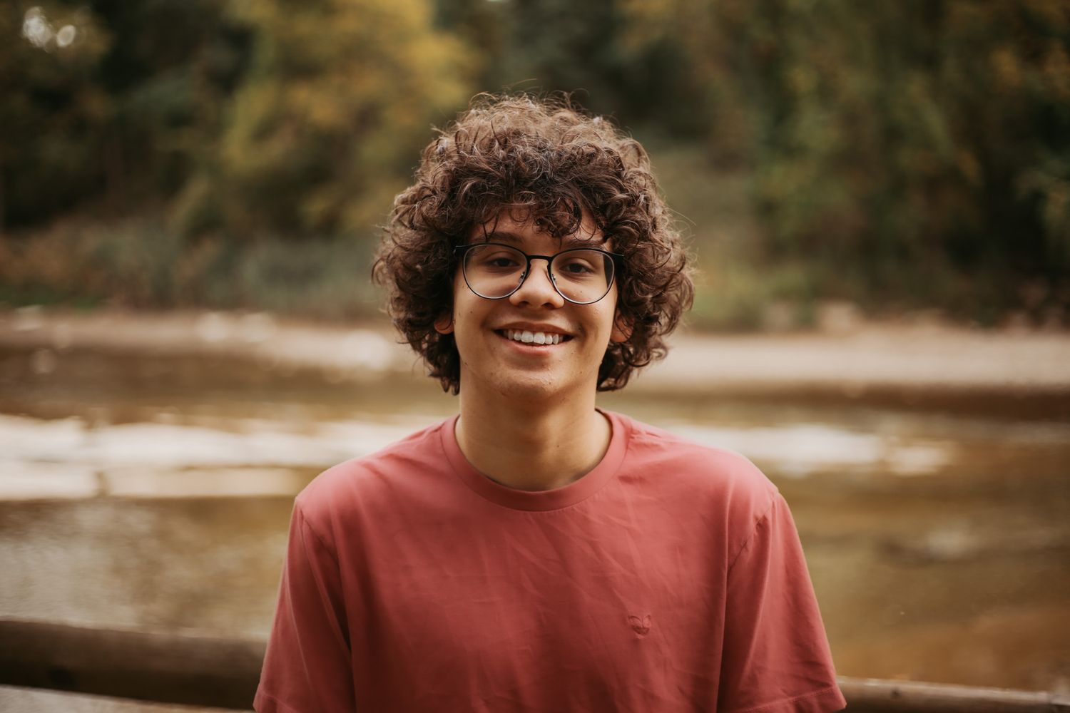 Person in red sweater smiling by a river on an autumn day.
