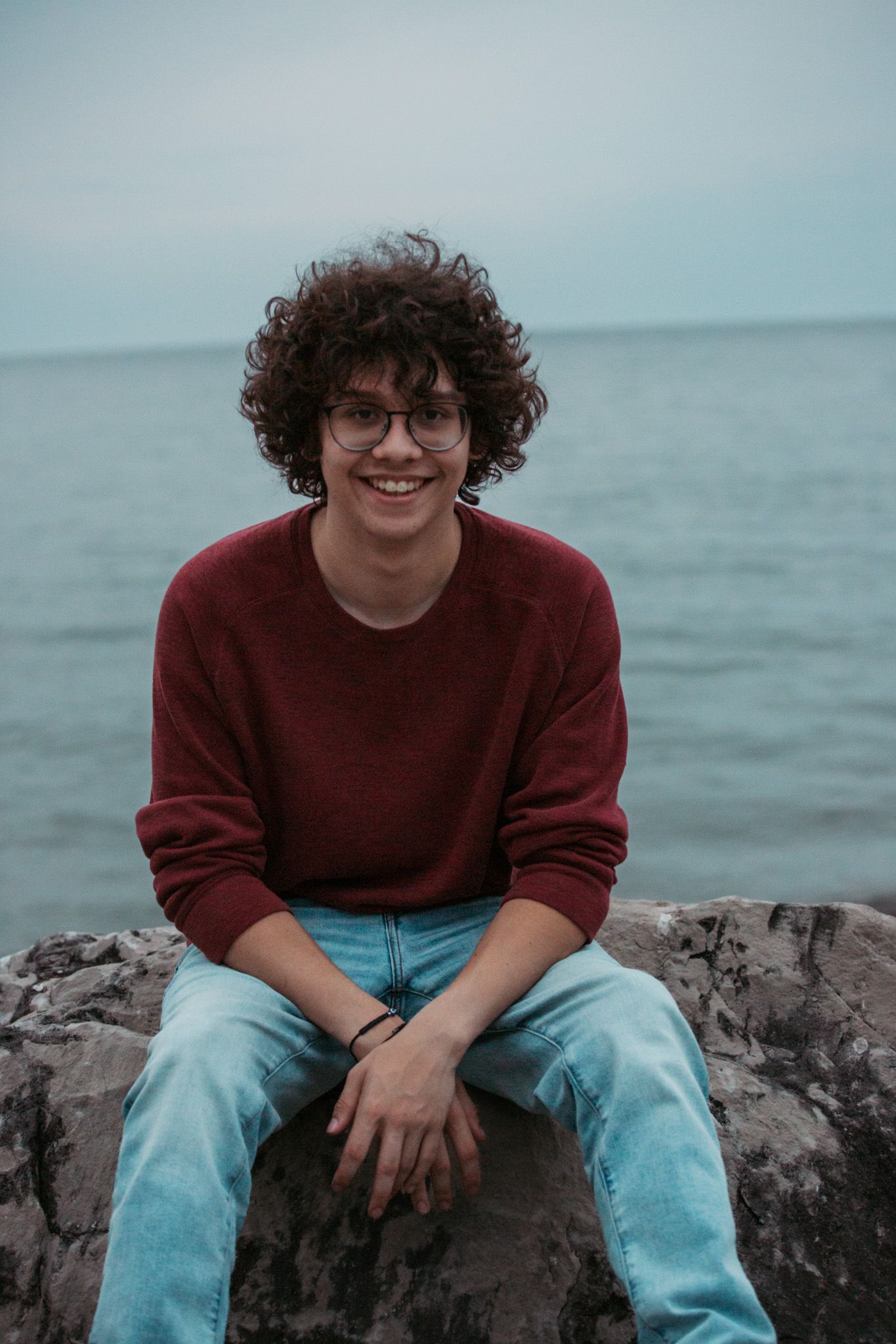 Person in red sweater sitting on coastal rocks with ocean in background.