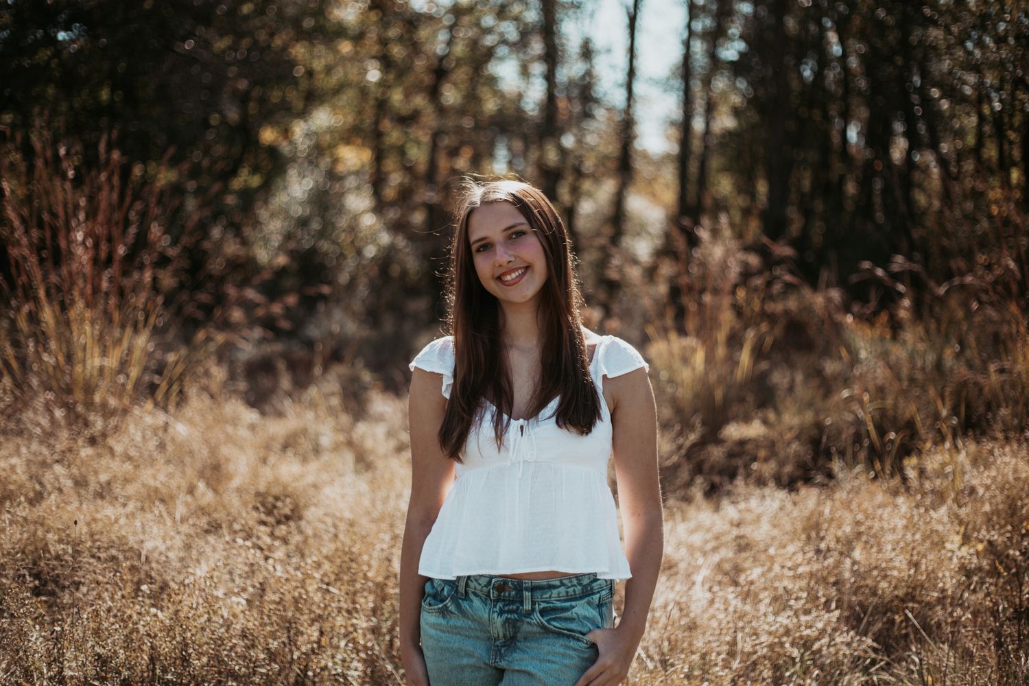 Casual outdoor portrait in white top and jeans against wooded background.