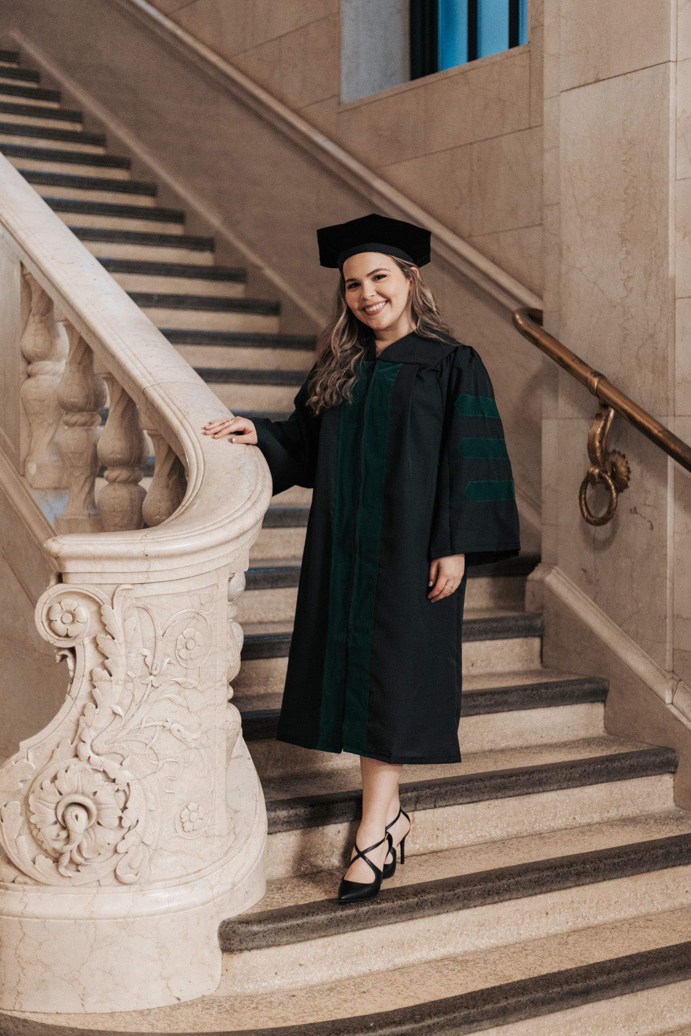 Graduate poses on ornate stone staircase in traditional black cap and gown.