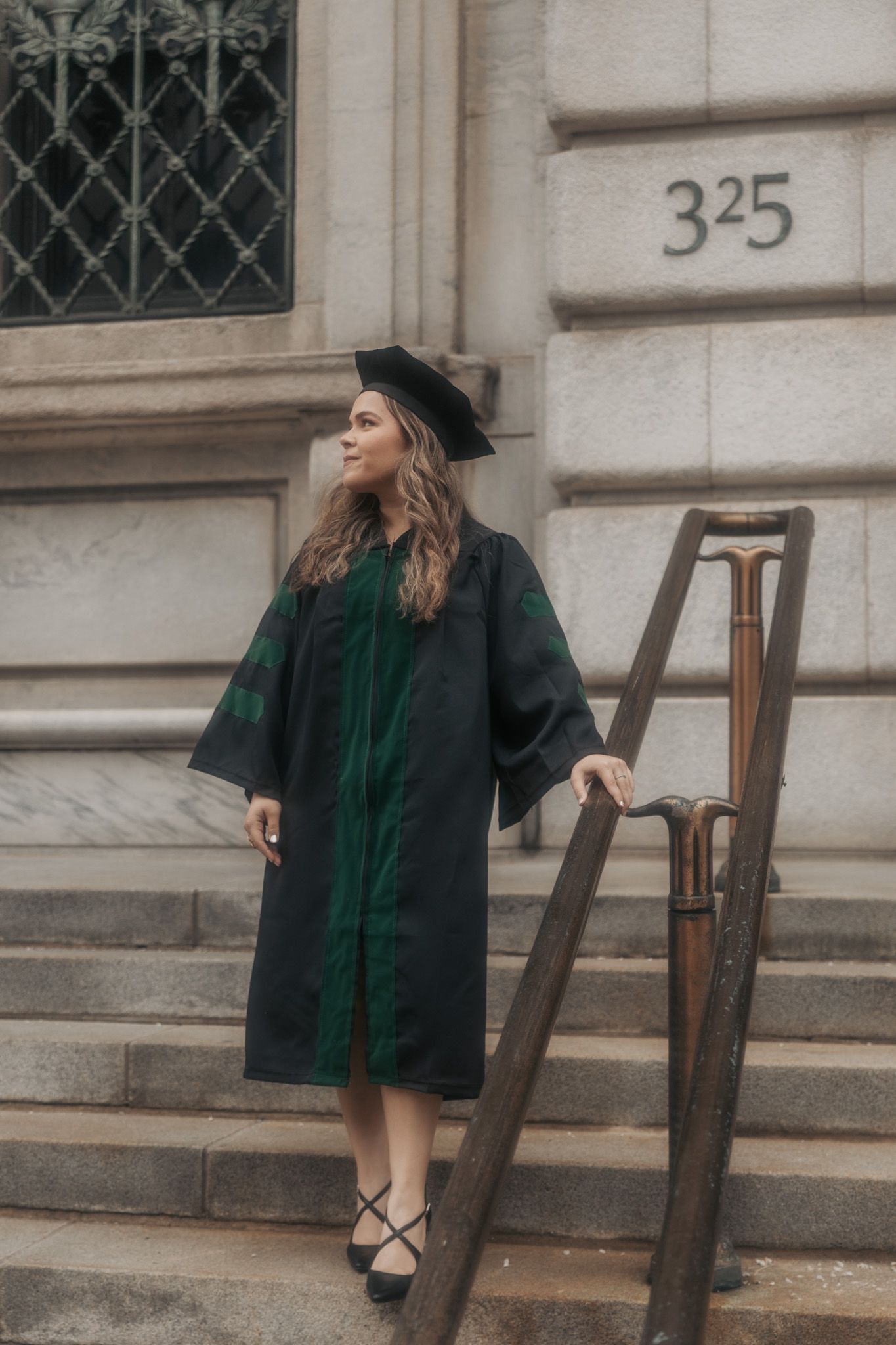 Graduate in dark academic regalia poses on historic building steps marked 325.