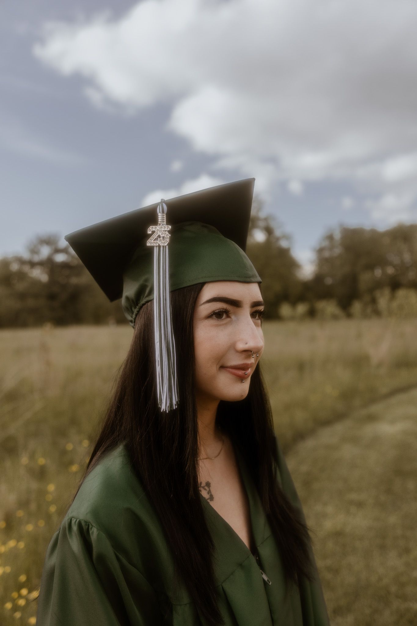 Graduate in dark green cap and gown poses in a sunny meadow with yellow wildflowers.
