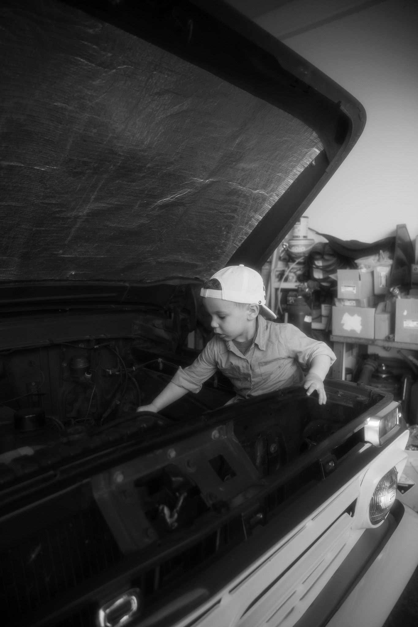Someone inspects under the hood of a vehicle in a black and white photograph.