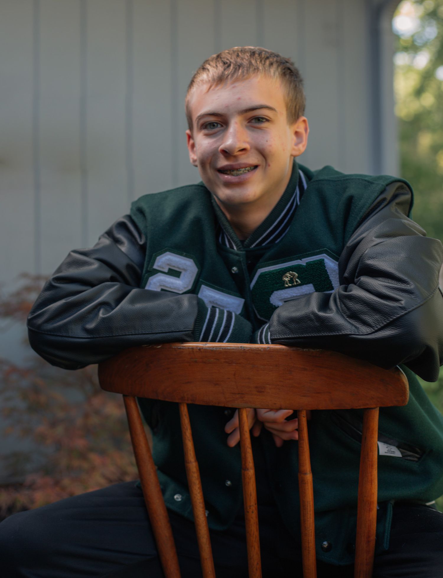 Person in varsity jacket leans casually on wooden chair in outdoor setting.