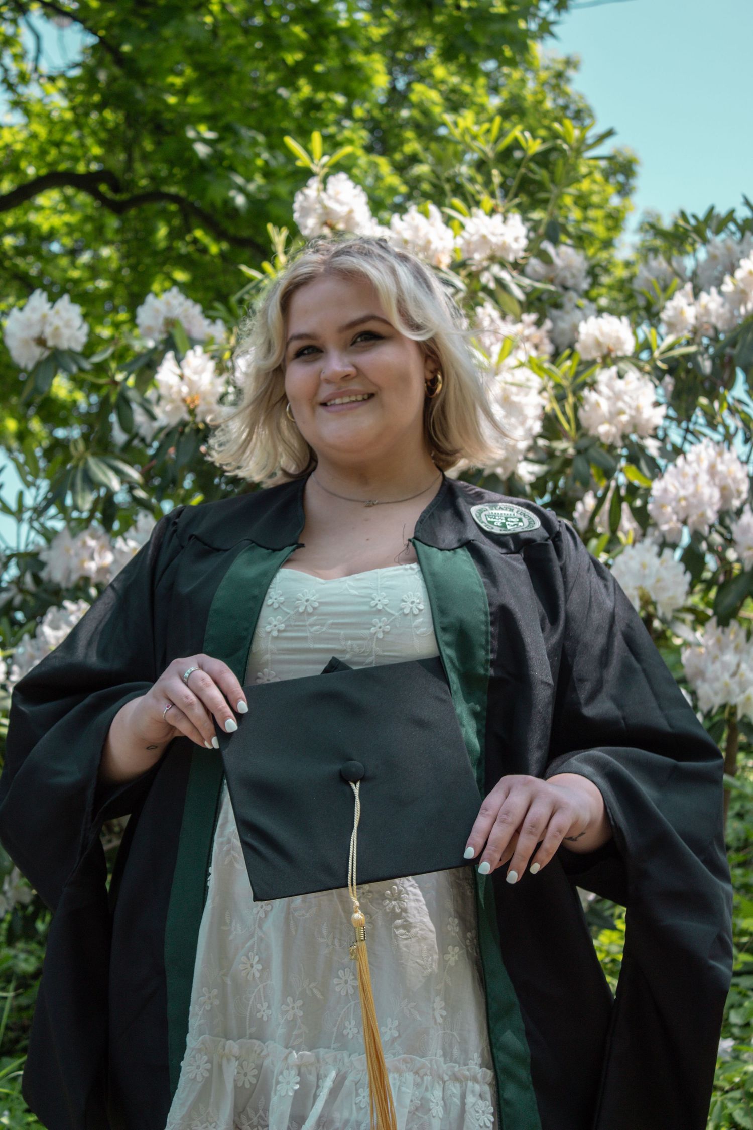 Graduate in black cap and gown poses in front of blooming white flowers on a sunny spring day.