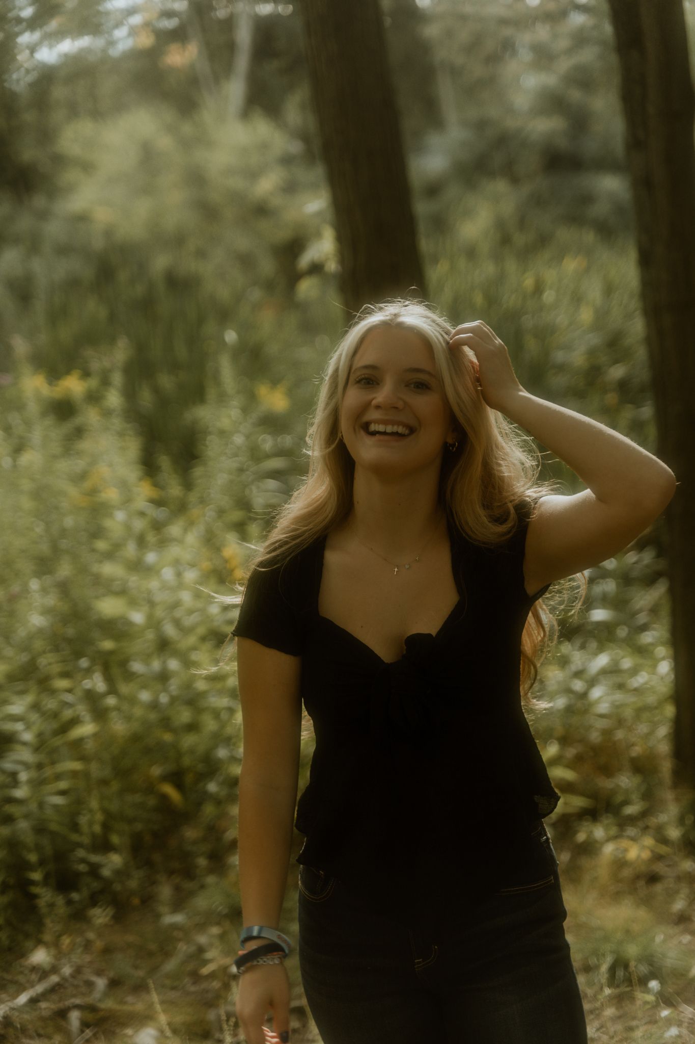 A woman in a black dress smiles joyfully while standing in a sunlit forest setting.
