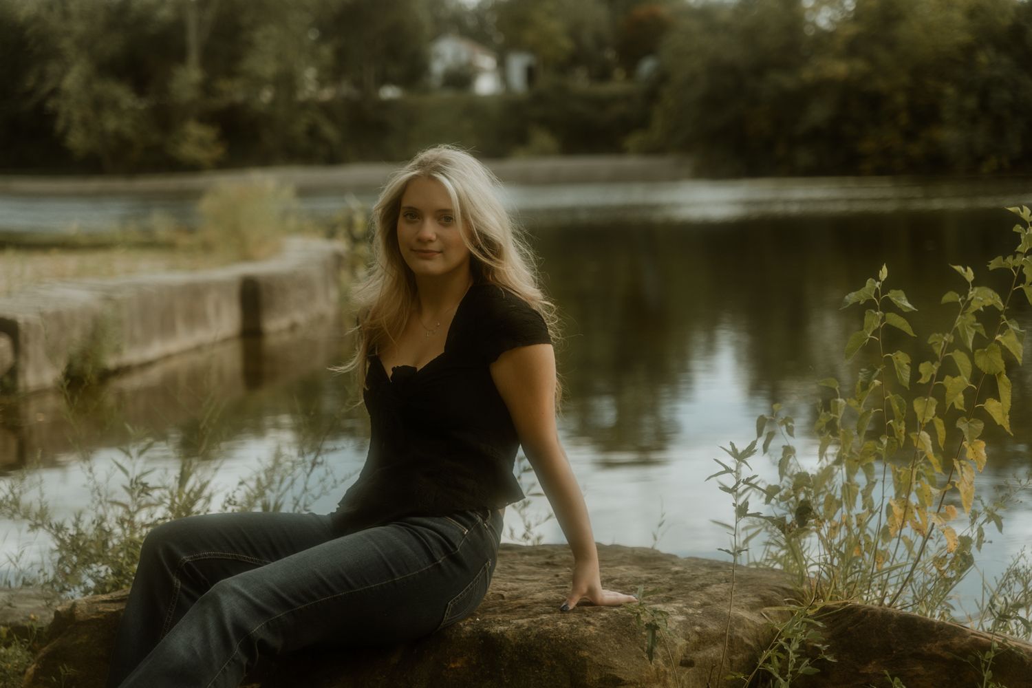 A woman in black dress sits by a serene pond on an autumn day.