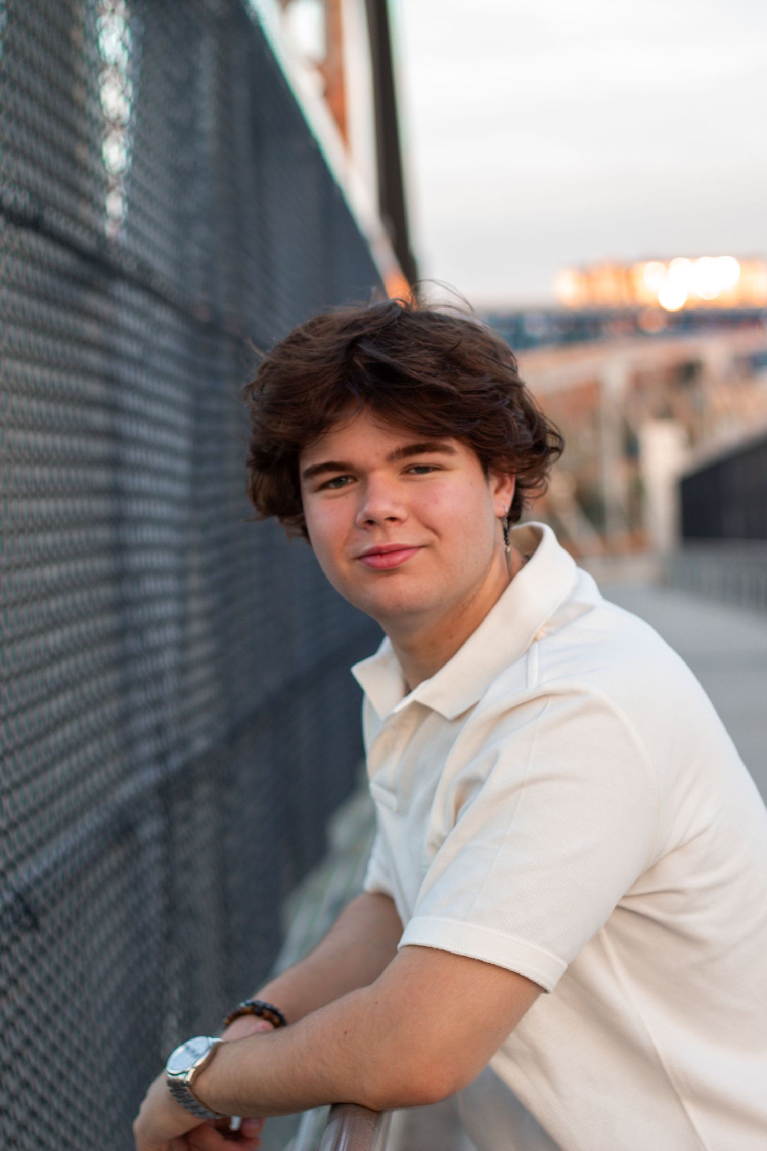 Person in white polo shirt leaning against metal fence during sunset in urban setting.
